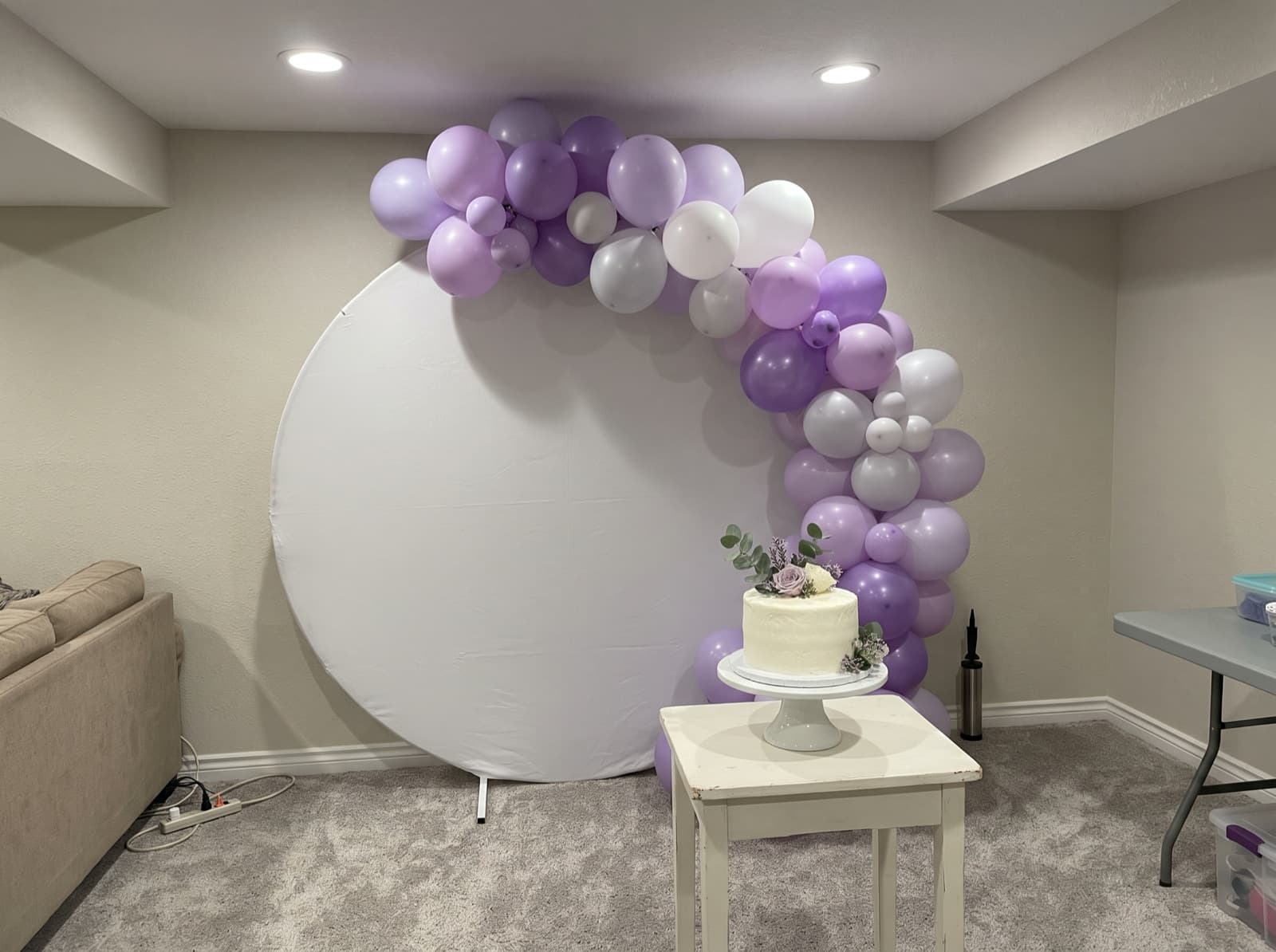 Round backdrop with a lavender and white organic balloon garland and a cake stand at a birthday party